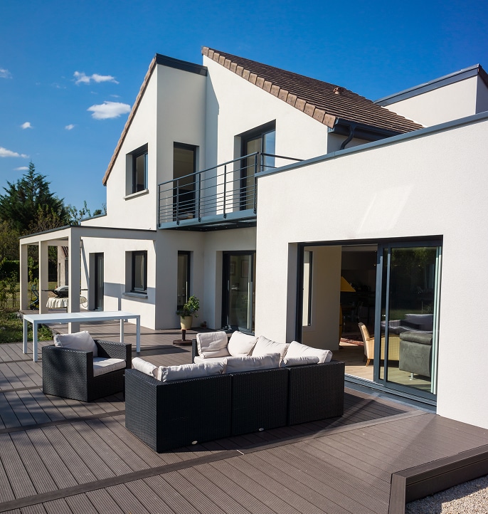 Maison moderne : terrasse composite et salon de jardin design. Maison moderne blanche, terrasse composite foncée, salon de jardin noir et baies vitrées. Balcon métallique à l'étage.