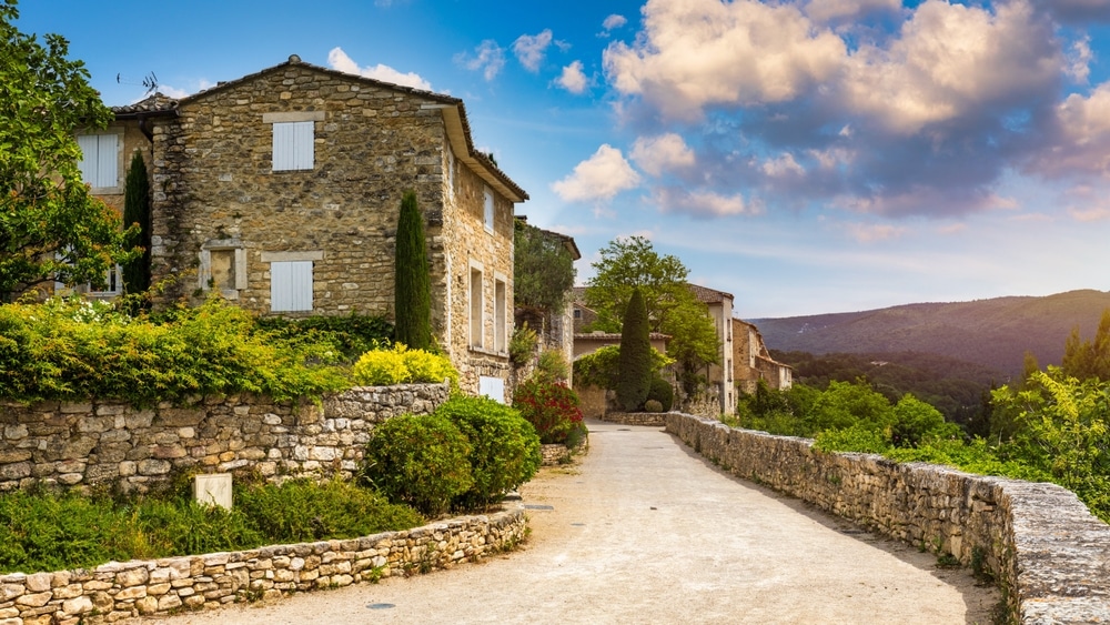 Ruelle ancienne en pierre de Provence avec vue idyllique. Ruelle en pierre d'un village provençal sous un ciel bleu. Vieilles maisons à volets blancs, murs de pierres sèches et cyprès. Vue sur la vallée.