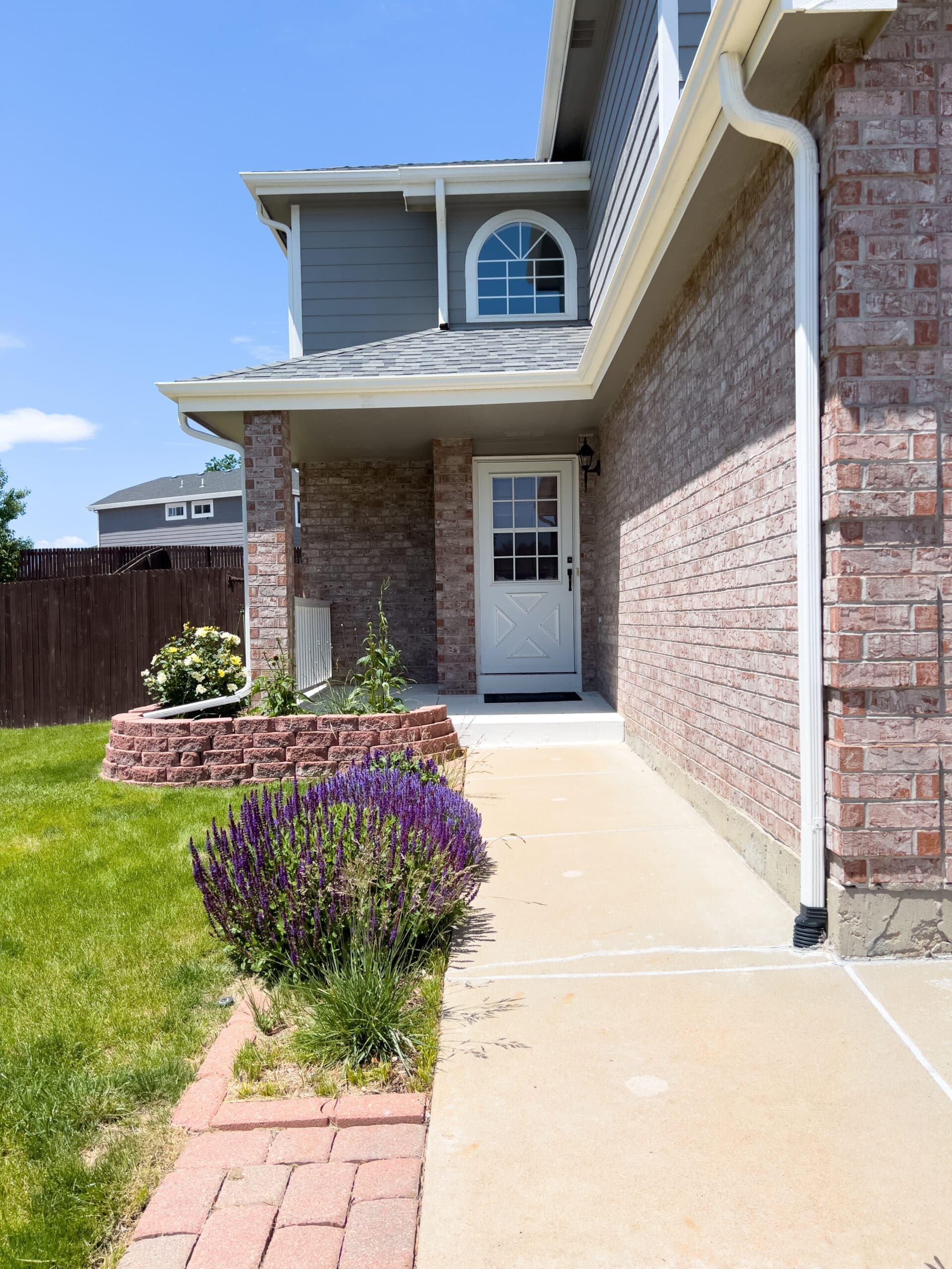 Entrée de Maison Moderne avec Briques et Jardin de Lavande Entrée de maison avec mur en briques, porte blanche, allée en béton et parterre de lavande violette sous un ciel bleu.
