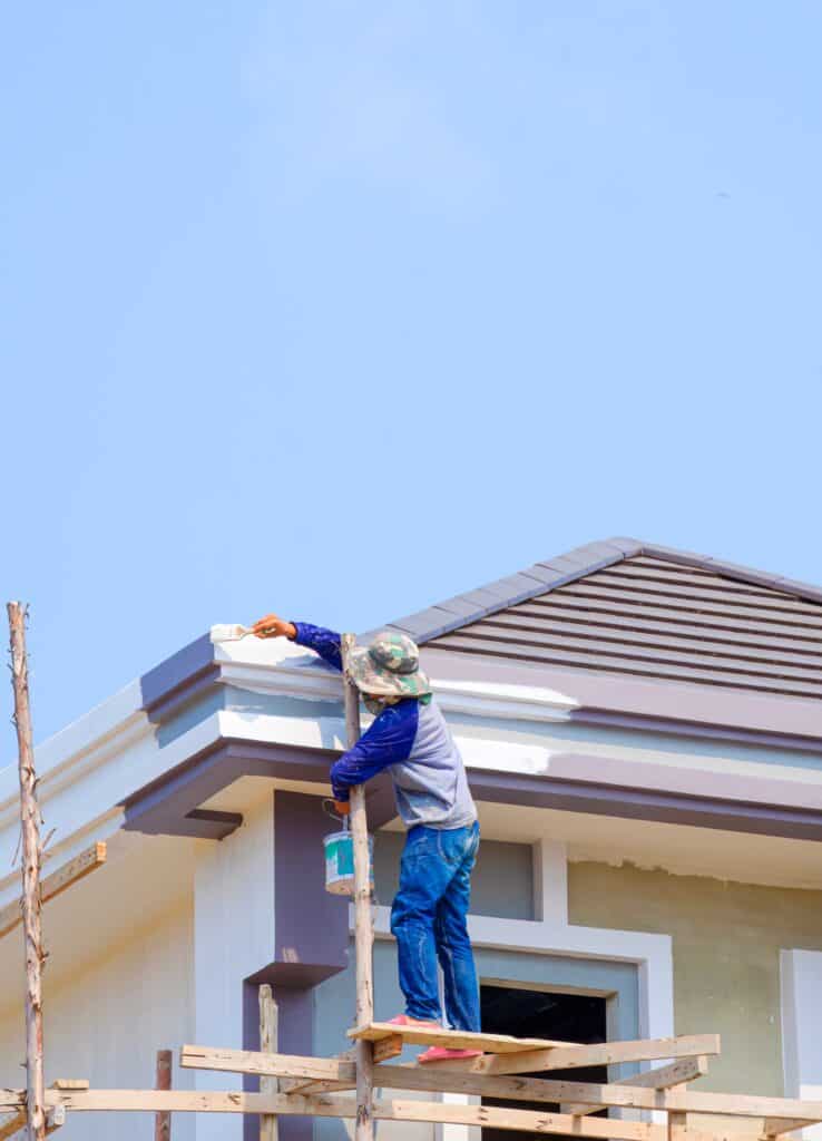 Travaux de peinture extérieure sur maison individuelle Peintre sur un échafaudage en bois peignant la façade d'une maison neuve sous un ciel bleu.